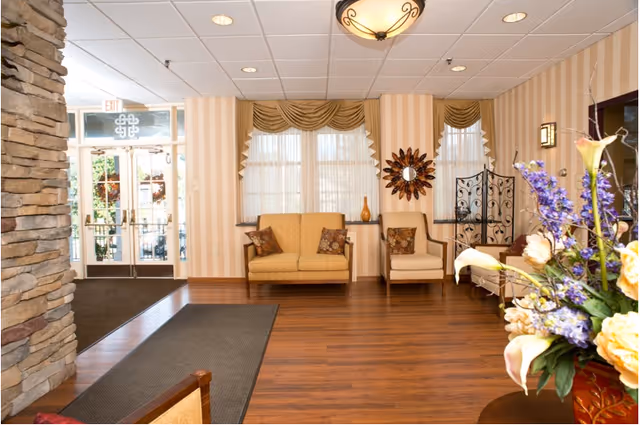 A bright and welcoming senior living facility lobby with wooden flooring, beige striped wallpaper, and large windows with sheer curtains and valances. There are two cushioned chairs with decorative pillows, a round mirror with a sunburst frame on the wall, a decorative metal room divider, and a floral arrangement with purple and yellow flowers on a table. The entrance door with glass panels is visible on the left side.
