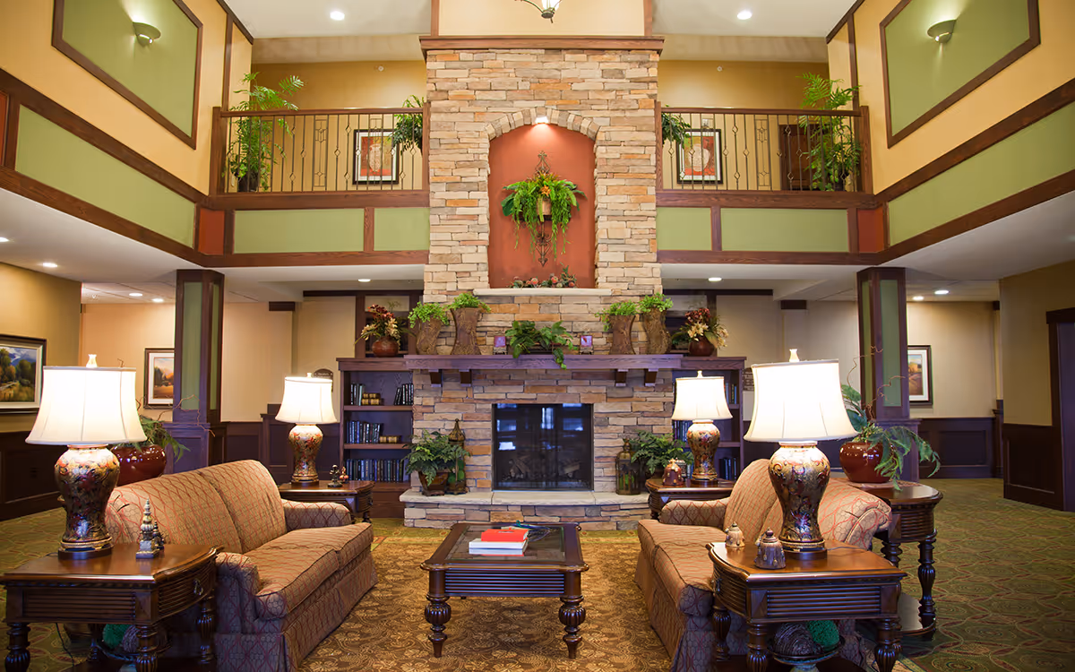 A spacious and warmly decorated senior living facility common area with two patterned sofas facing each other, a wooden coffee table with books, and a large stone fireplace adorned with plants and decorative items. The room features a high ceiling with a balcony railing above, green and beige walls with wood trim, and several table lamps providing soft lighting.