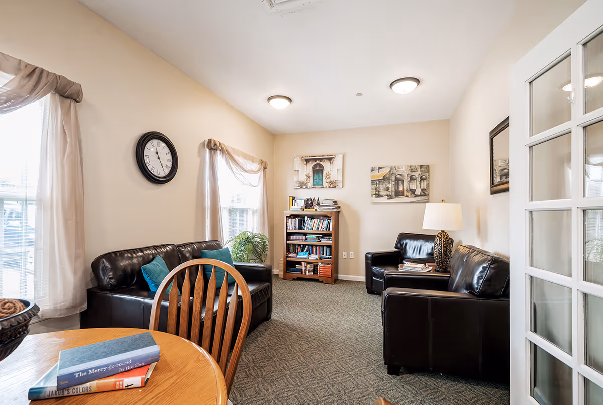 Cozy common room with leather sofas, a bookcase, round wooden table and chairs, wall art, and windows with sheer curtains.