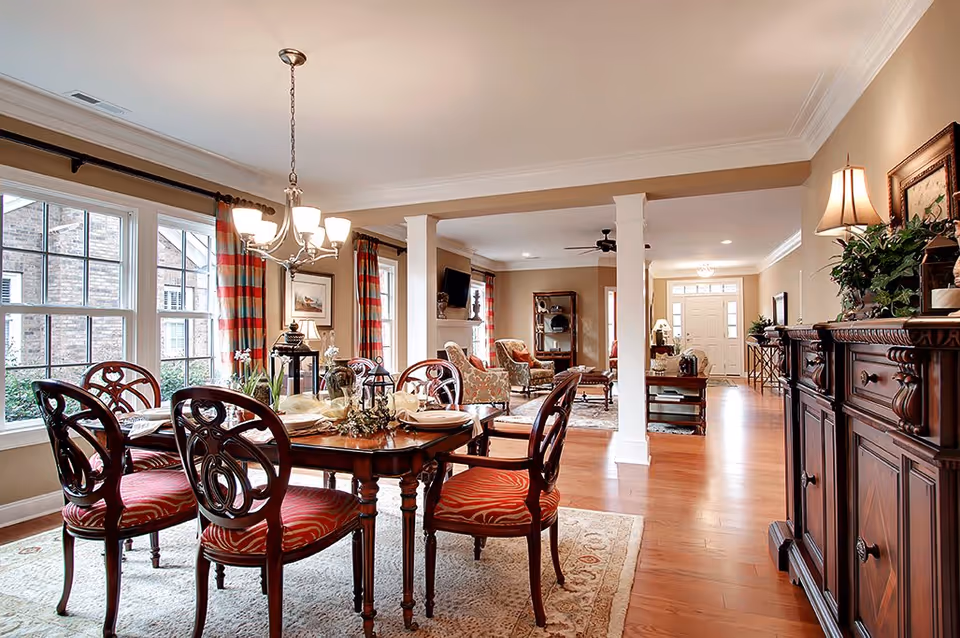 A spacious and elegant interior featuring a dining area with a wooden table and six chairs with red patterned cushions. The dining area is illuminated by a chandelier and has large windows with red and beige striped curtains. Beyond the dining area is a living room with upholstered armchairs, a coffee table, a wall-mounted TV, and a ceiling fan. The room has hardwood floors, beige walls, and decorative furniture including a sideboard with plants and framed artwork.