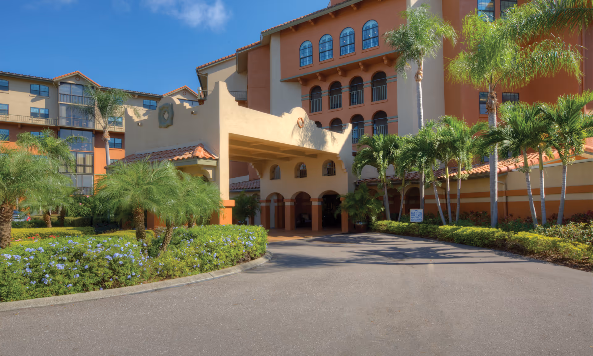 Exterior view of a multi-story senior living facility with a covered entrance, surrounded by palm trees and well-maintained landscaping under a clear blue sky.