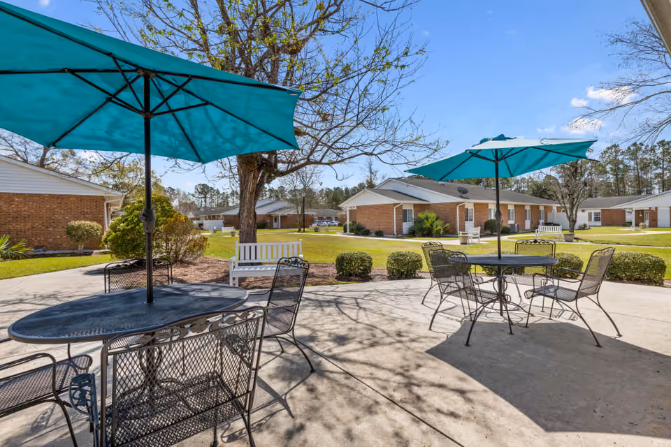 Outdoor patio area with two black metal tables, each shaded by a teal umbrella. Each table is surrounded by black metal chairs. The patio overlooks a grassy courtyard with bushes, trees, and single-story brick buildings in the background under a clear blue sky.
