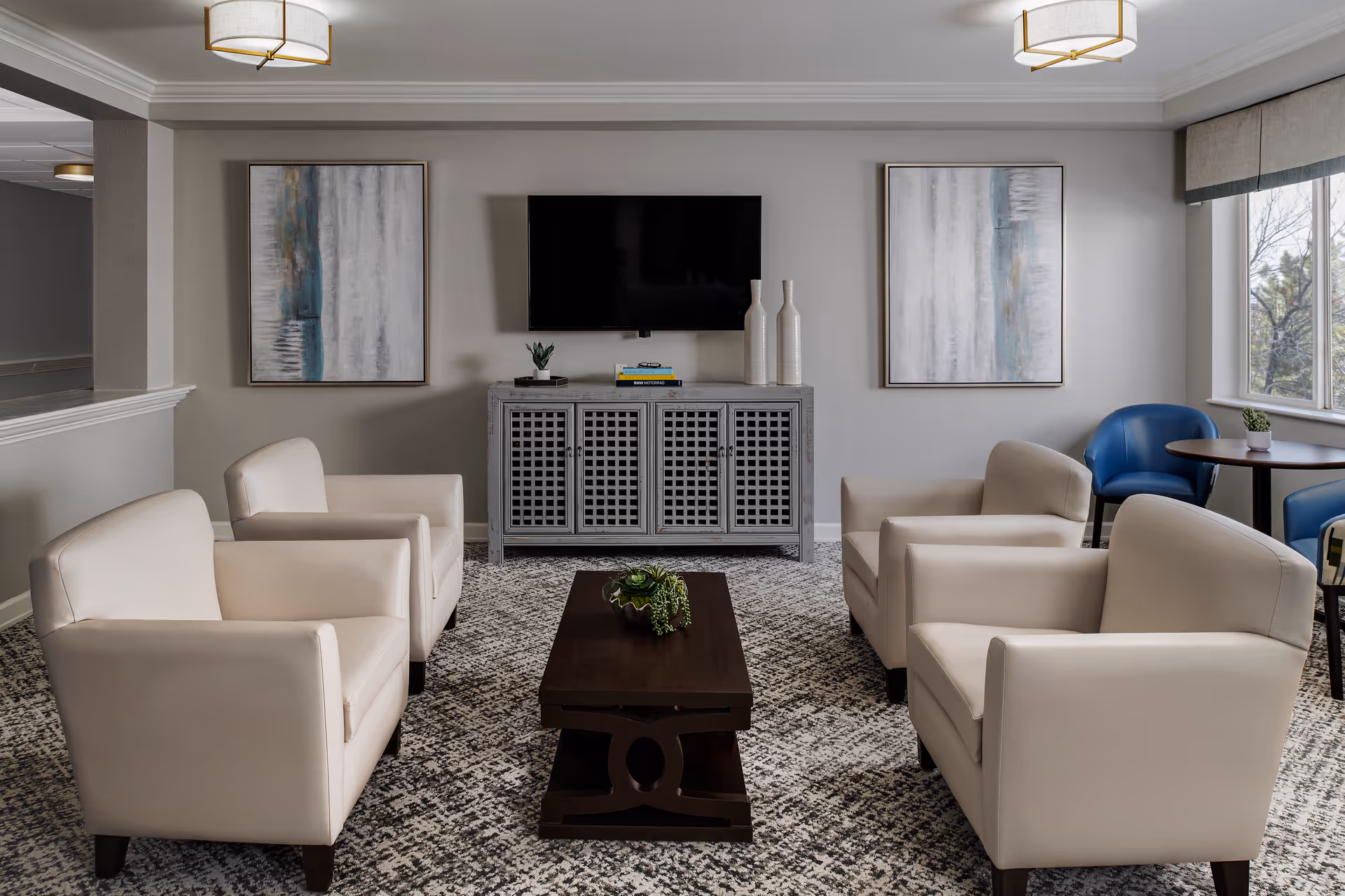A modern living room with four beige armchairs arranged around a dark wooden coffee table with a small plant. A gray cabinet with a flat-screen TV mounted above it is against the wall, flanked by two abstract paintings. Two blue chairs and a small round table are near a window on the right side, letting in natural light.