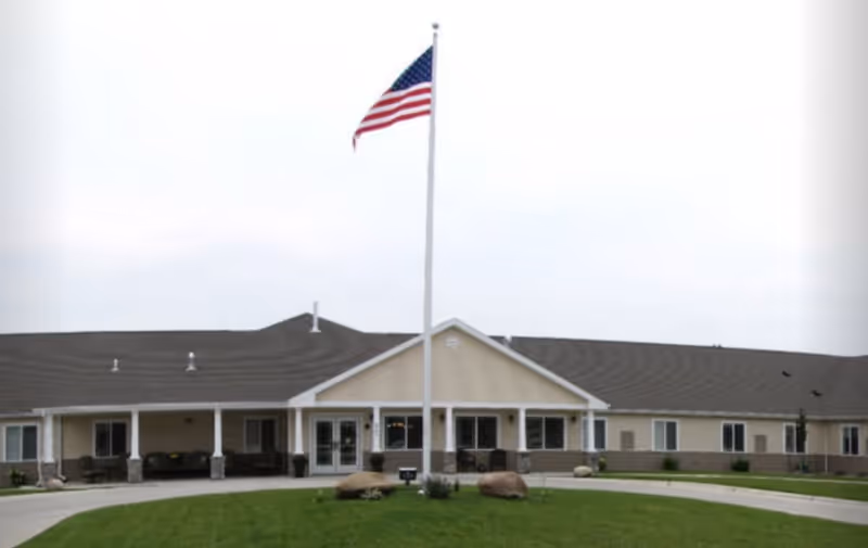 Exterior view of a single-story senior living facility building with a large American flag on a flagpole in front. The building has a covered entrance with seating areas and a circular driveway surrounded by green grass and landscaping.