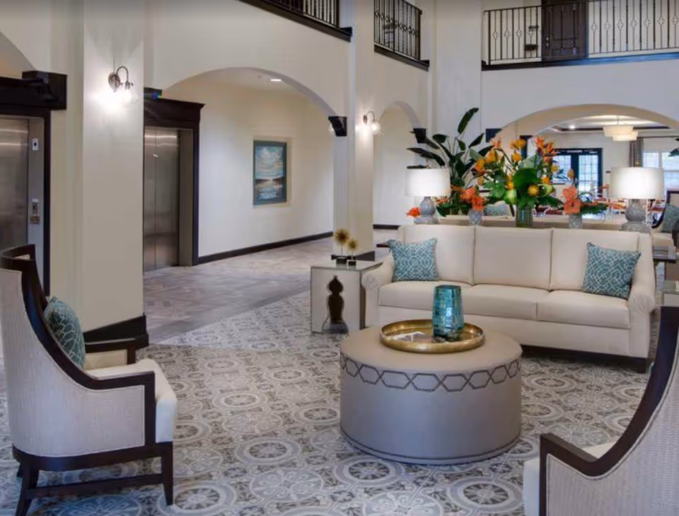 Spacious furnished lounge with sofas, chairs, an ottoman, decorative plants and elevators visible in the background.