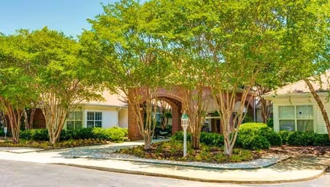 Exterior view of a single-story assisted living facility with brick and white siding, surrounded by green trees and shrubs under a clear sky.