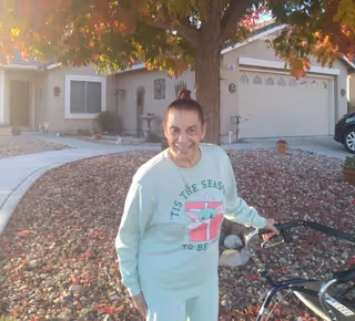 An elderly woman smiling outdoors in front of a residential house with a tree and autumn leaves. She is wearing a light-colored sweatshirt with a holiday-themed design and is holding a walker.