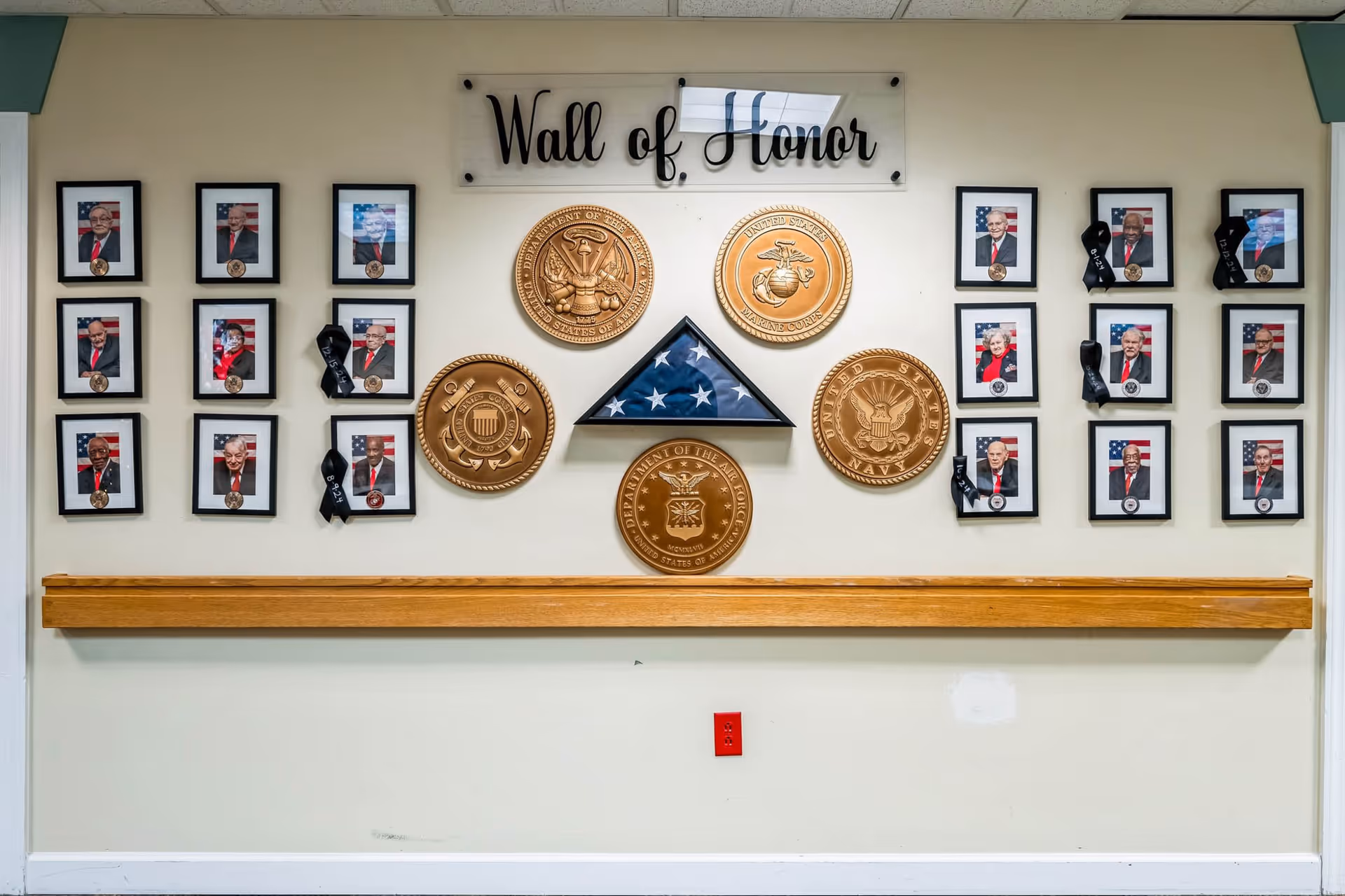 Wall of Honor display featuring framed photographs of veterans with black ribbons, five large bronze military emblems representing different branches of the U.S. Armed Forces, and a folded American flag in a triangular case mounted on a white wall above a wooden handrail.