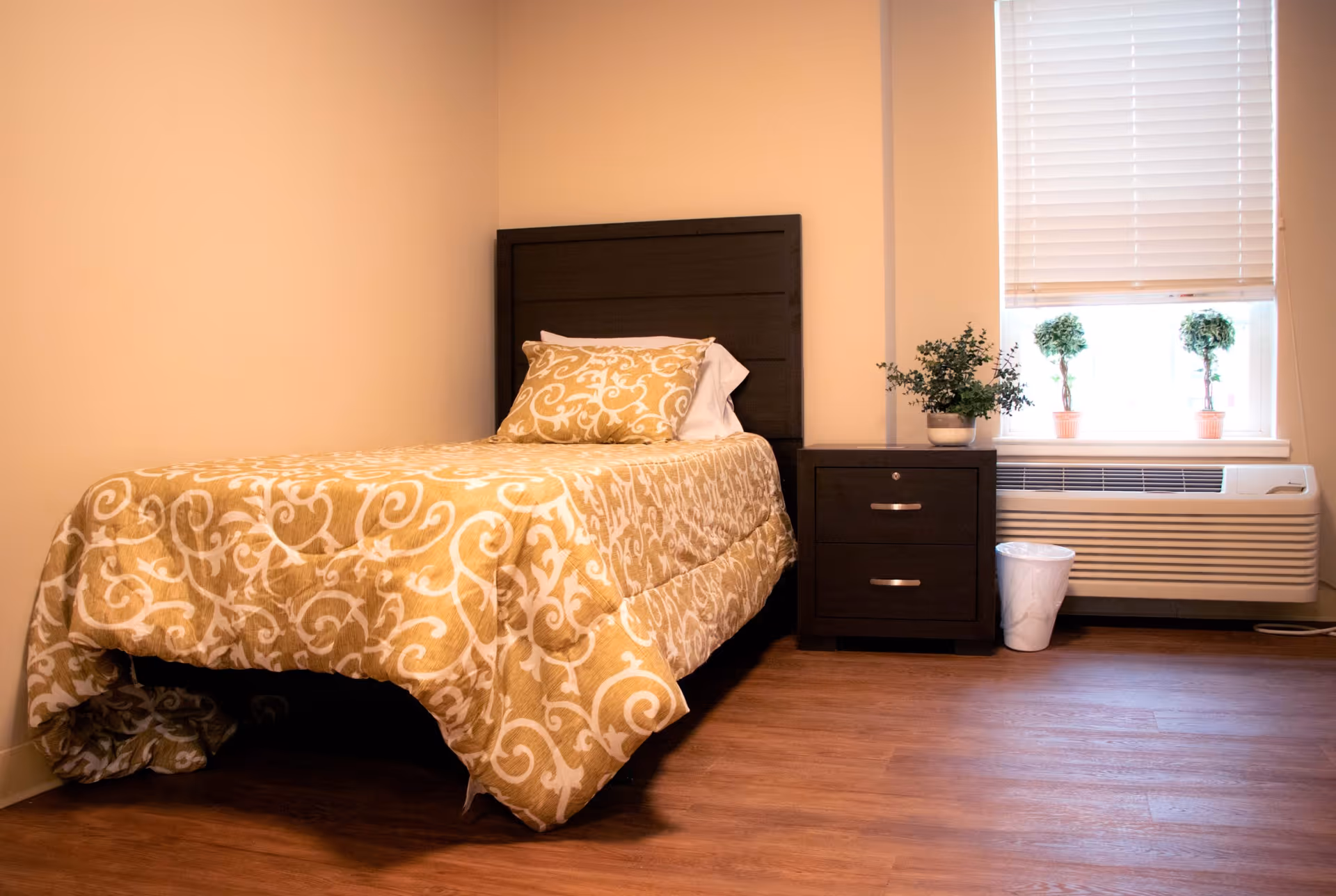 Simple furnished bedroom with a single bed, patterned bedding, a nightstand, potted plants on the windowsill, and an air conditioner unit.