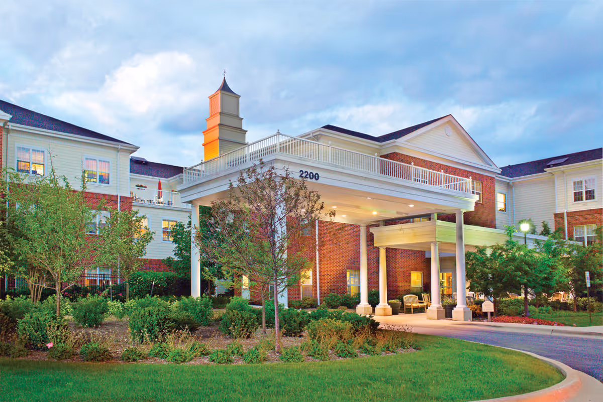 Exterior view of Belmont Village Senior Living Glenview building at dusk, showing a large covered entrance with white columns, well-maintained landscaping with bushes and trees, and a multi-story brick and siding facade with lit windows.