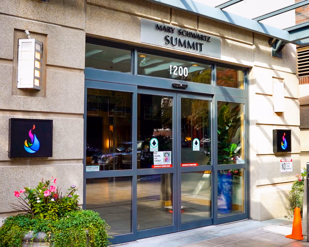 Entrance to the Mary Schwartz Summit facility featuring glass automatic sliding doors with the address number 1200 above. The building exterior is made of stone with two colorful flame logos on either side of the entrance and potted plants nearby.
