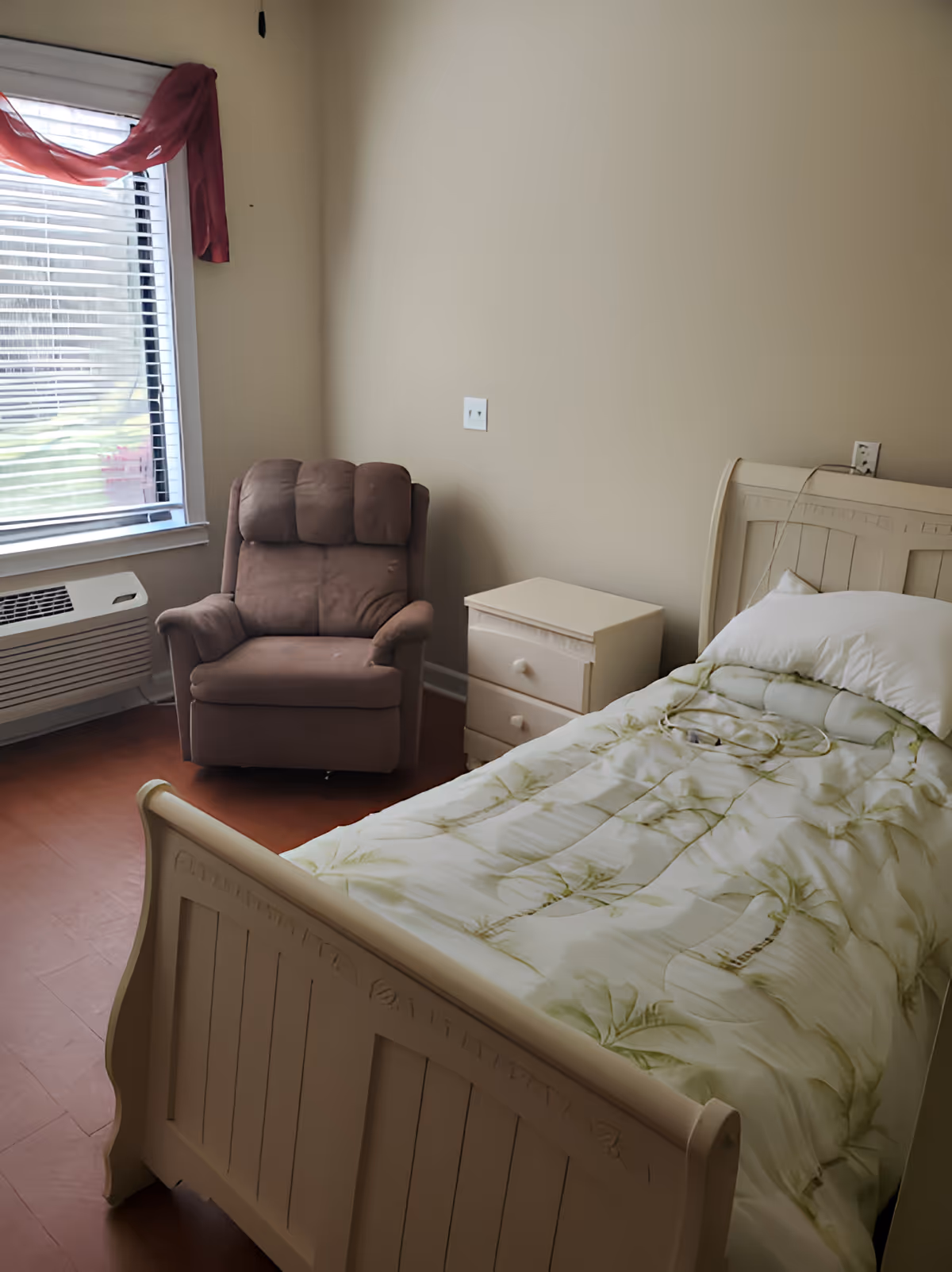 A small bedroom with a single bed covered with a light-colored blanket featuring a green leaf pattern. Next to the bed is a white nightstand with two drawers. A brown cushioned armchair is placed near a window with blinds and a red curtain draped over the top. The room has beige walls and a reddish-brown floor.