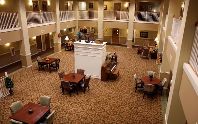 Spacious senior living facility common area with patterned carpet, multiple seating arrangements including tables and chairs, a piano, and a central white column. The room has two levels with white railings on the upper level and warm lighting on the walls.