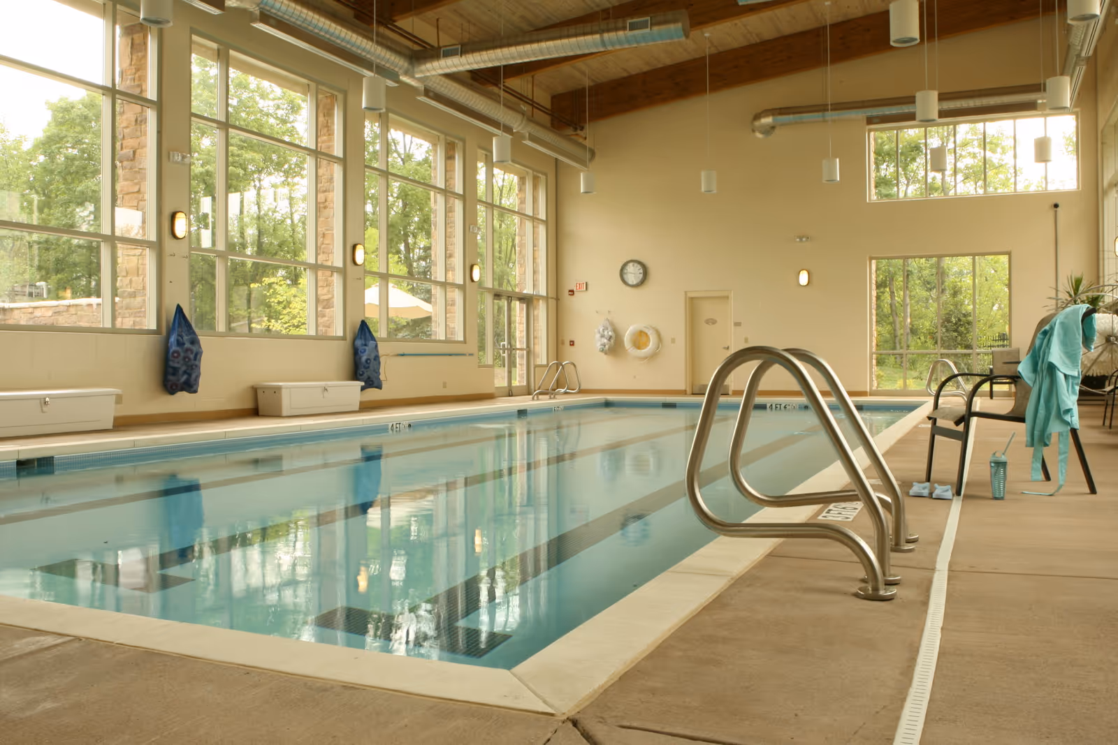 Indoor swimming pool with large windows letting in natural light. The pool has metal handrails and lane markings. There are chairs and a towel on the right side, with a water bottle and slippers nearby. The walls are light-colored with a clock and safety equipment mounted on one wall.