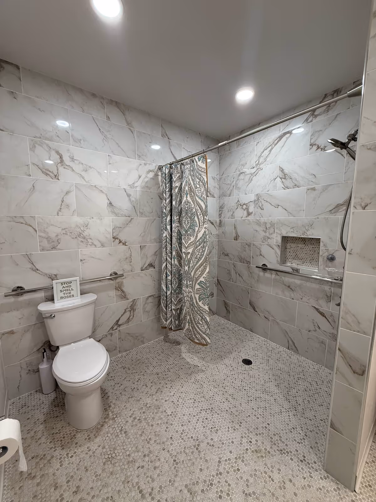 A modern bathroom with marble-patterned wall tiles and small circular mosaic floor tiles. The bathroom features a white toilet with a sign on top that reads 'STOP AND SMELL THE ROSES'. There is a shower area with a patterned shower curtain, a handheld showerhead, and a built-in niche in the wall. The room is well-lit with recessed ceiling lights.
