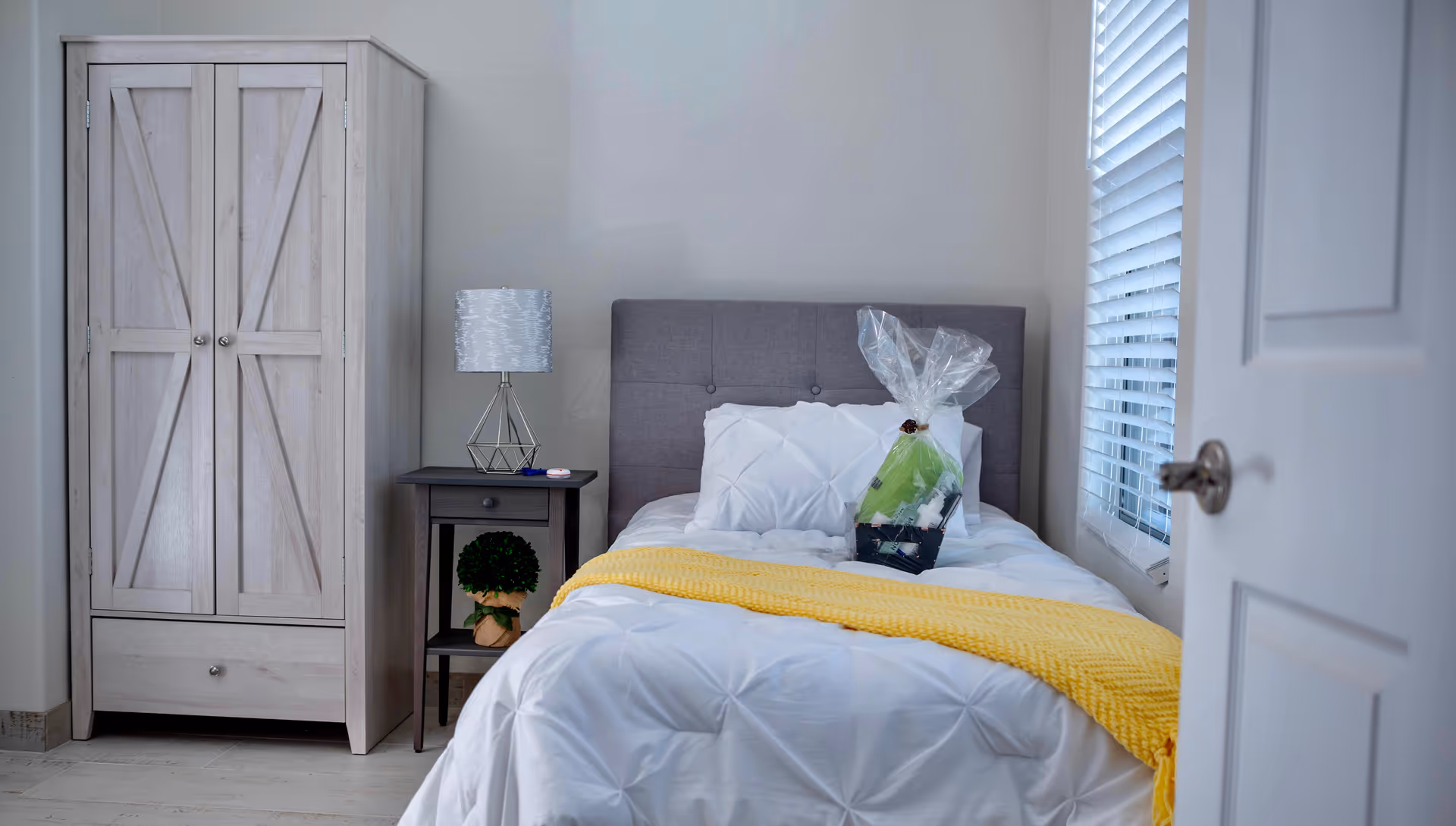 A small bedroom with a single bed featuring a gray upholstered headboard, white bedding, and a yellow throw blanket. On the bed is a gift basket wrapped in clear plastic. Next to the bed is a dark wooden nightstand with a modern lamp and a small potted plant underneath. To the left of the nightstand is a light wood wardrobe. A window with white blinds is on the right wall, and a partially open white door is visible on the right side of the image.