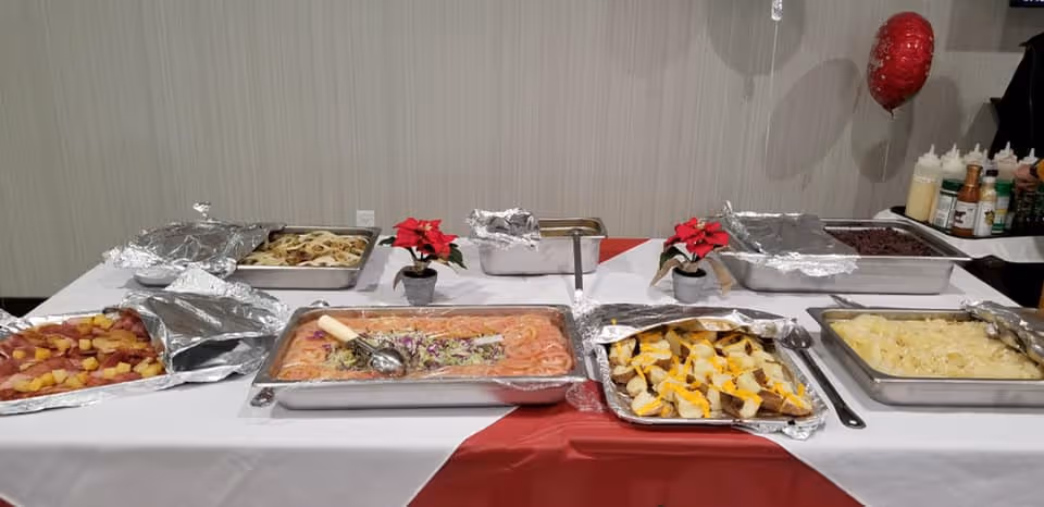 A buffet table with various trays of food covered partially with aluminum foil, including pasta, vegetables, and other dishes. The table is covered with a white and red tablecloth and decorated with small red poinsettia plants. Condiment bottles and a red balloon are visible in the background.