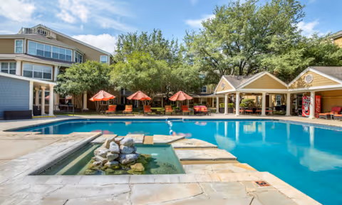 Outdoor swimming pool area at Independence Hill Retirement Community with a small rock water feature in the foreground, surrounded by lounge chairs with red umbrellas, trees, and a covered pavilion with a Coca-Cola vending machine.