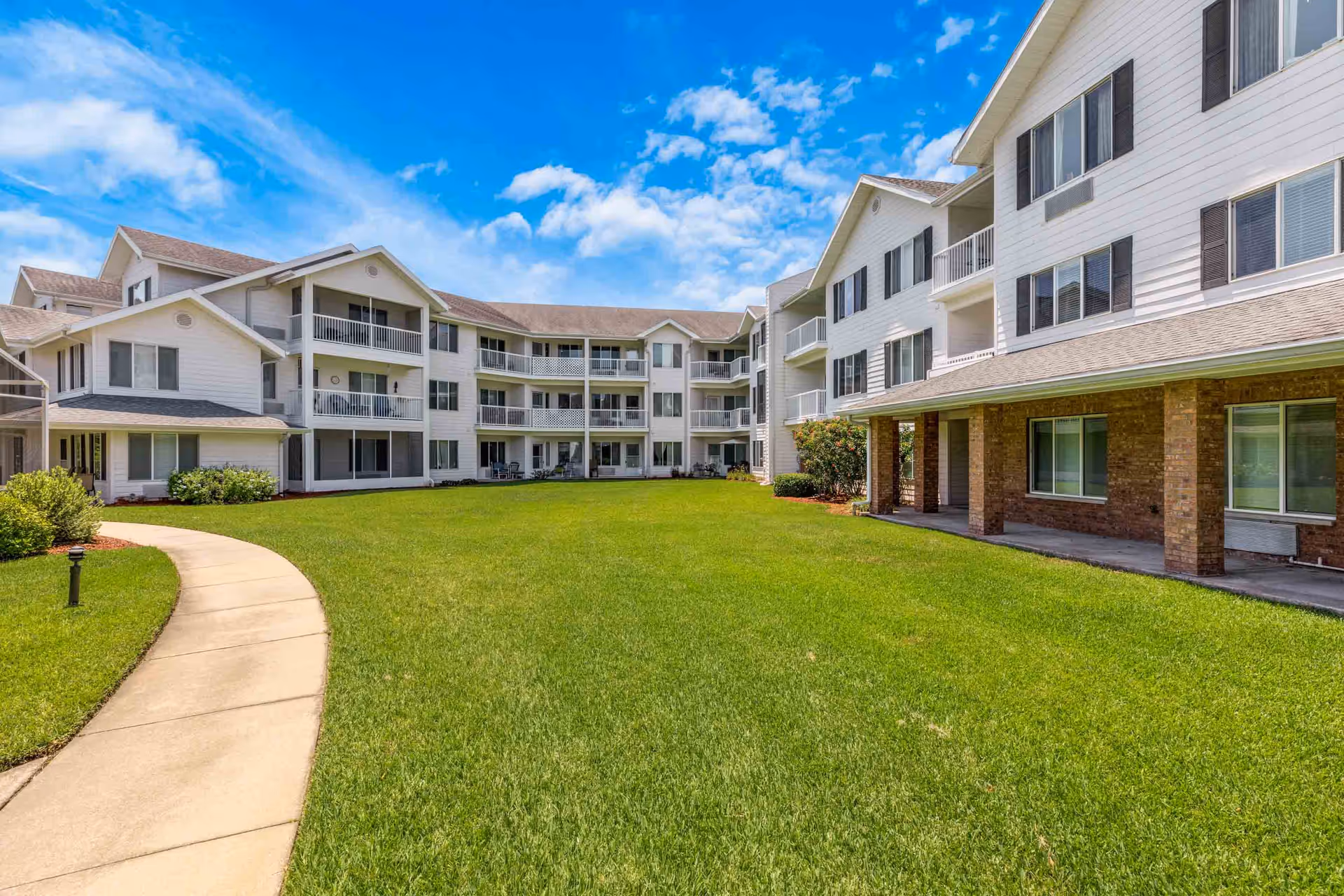 Exterior view of The Tremont - A Provincial Senior Living Community showing a large green lawn with a curved concrete pathway leading towards a multi-story white building with balconies and windows under a partly cloudy blue sky.