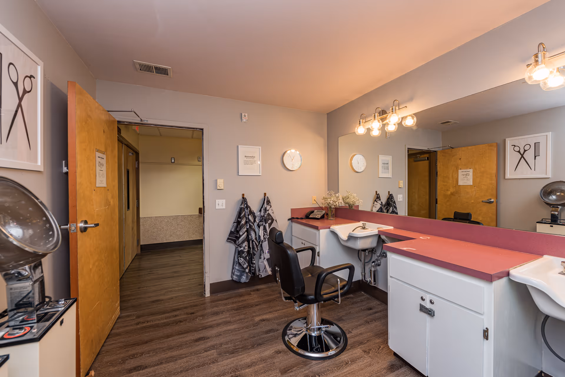 Interior view of a hair salon area in a senior living facility with a styling chair, two sinks, a large mirror, hair drying equipment, and hair cutting capes hanging on hooks. The room has wooden flooring, beige walls, and bright lighting fixtures above the mirror.