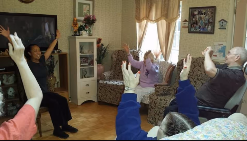 A group of residents and a caregiver seated in a living room raising their arms during a group exercise.