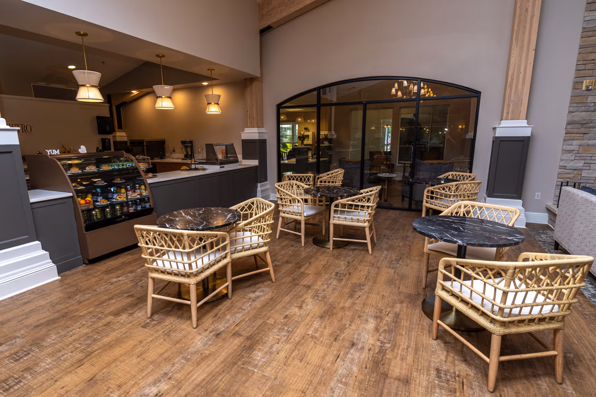 Interior view of a dining area with several round tables and wicker chairs with cushions. There is a counter with a display case containing food items and hanging pendant lights above. The floor is wooden, and there is a large window with a black frame showing another room with seating.