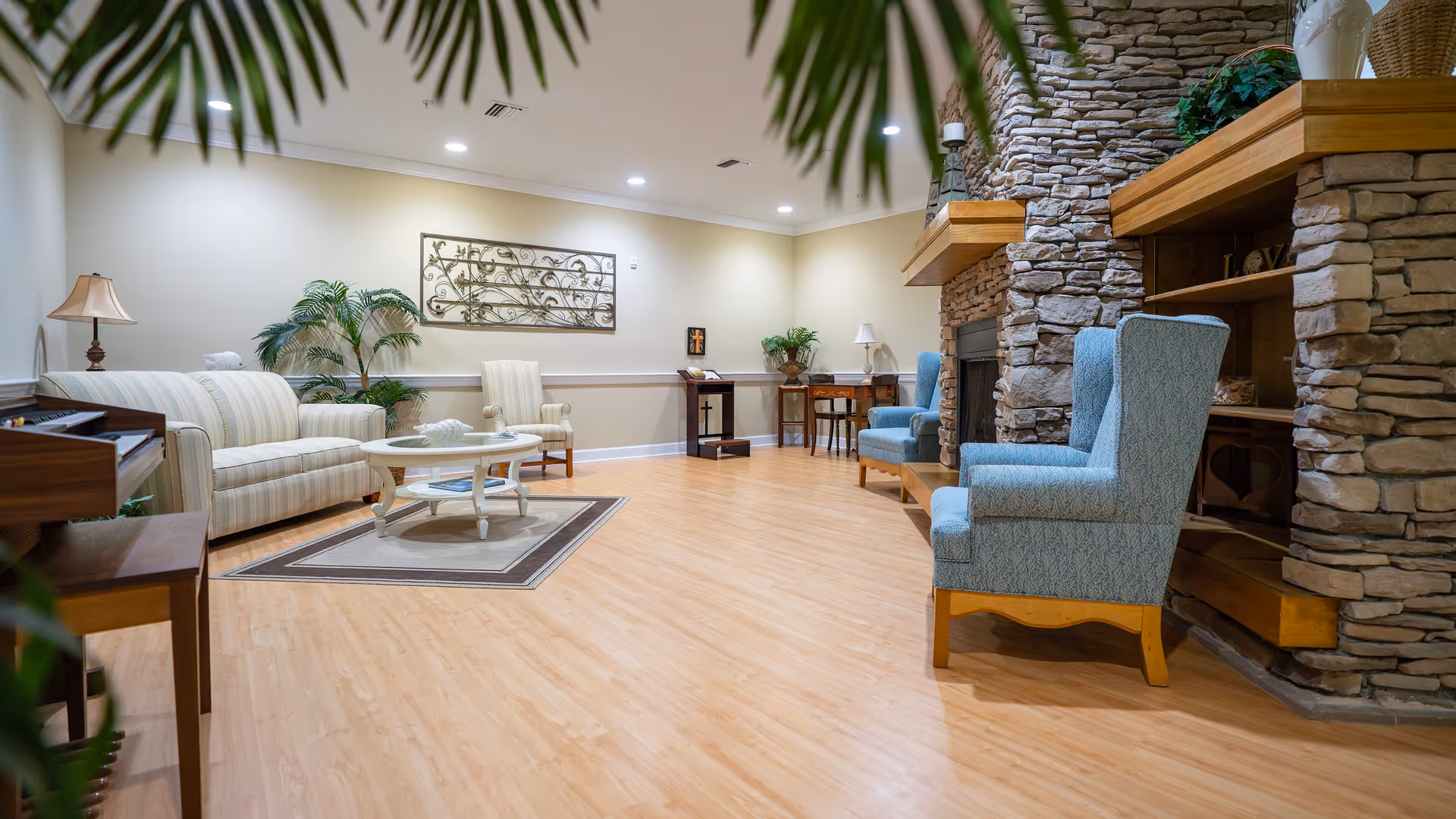 A cozy living room area with light wood flooring, a stone fireplace on the right, two blue upholstered armchairs, a striped sofa, a white round coffee table on a rug, a wooden piano, and decorative plants and wall art.