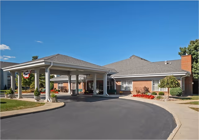 Exterior view of a single-story brick building with a covered entrance driveway, landscaped with shrubs and flowers under a clear blue sky.