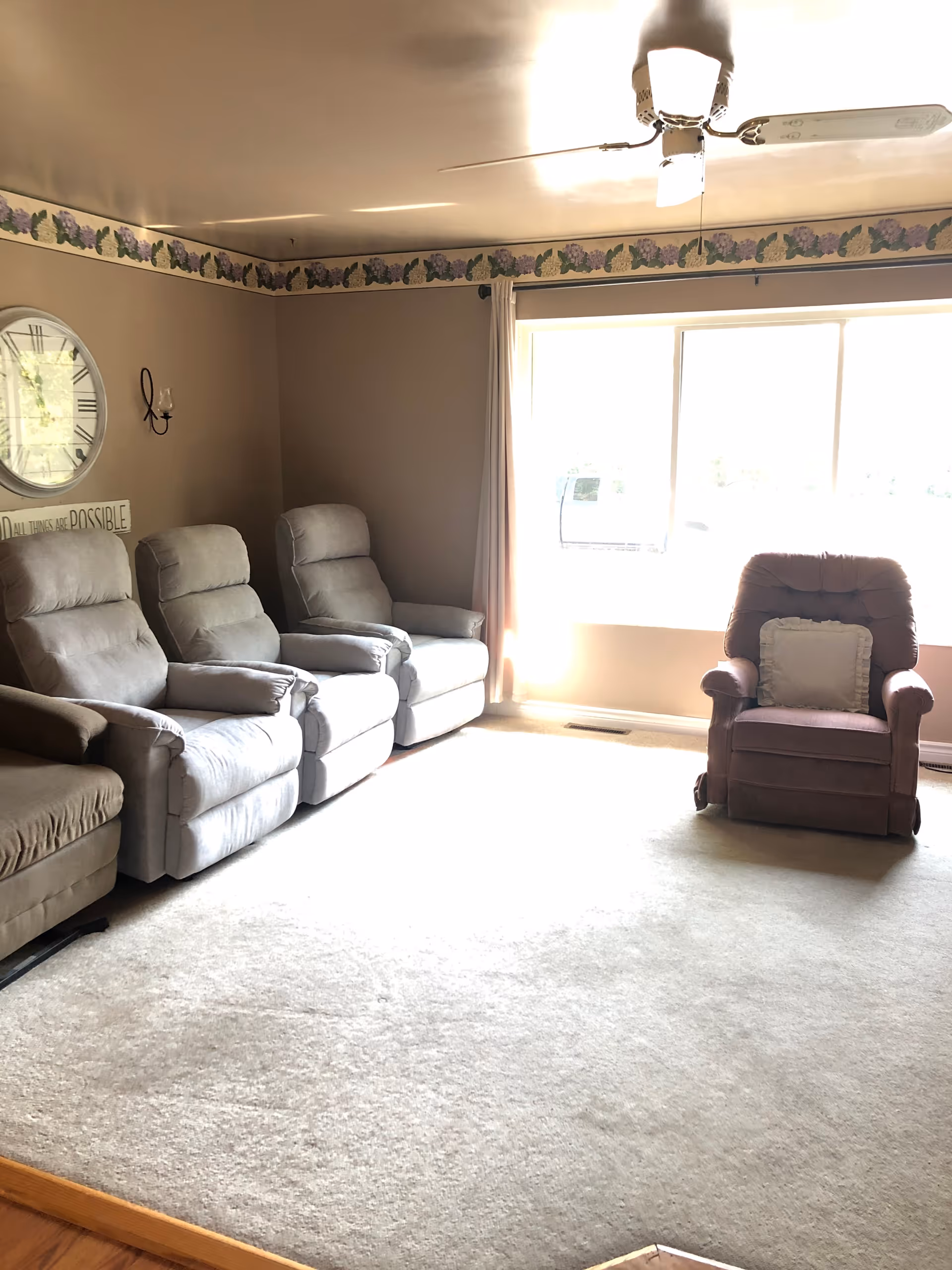 A living room with four recliner chairs arranged along the walls. Three light gray recliners are on the left side, and one maroon recliner with a pillow is near a large window with white curtains. The walls are beige with a floral border near the ceiling, and there is a ceiling fan with a light fixture. A large round clock and a wall decoration are visible on the left wall.