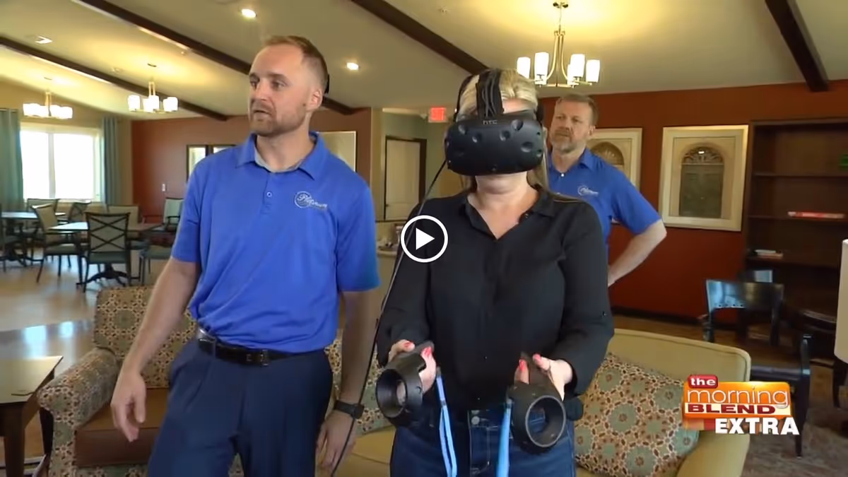 A woman wearing a virtual reality headset holds controllers in a cozy assisted living common room while two staff members in blue shirts look on.