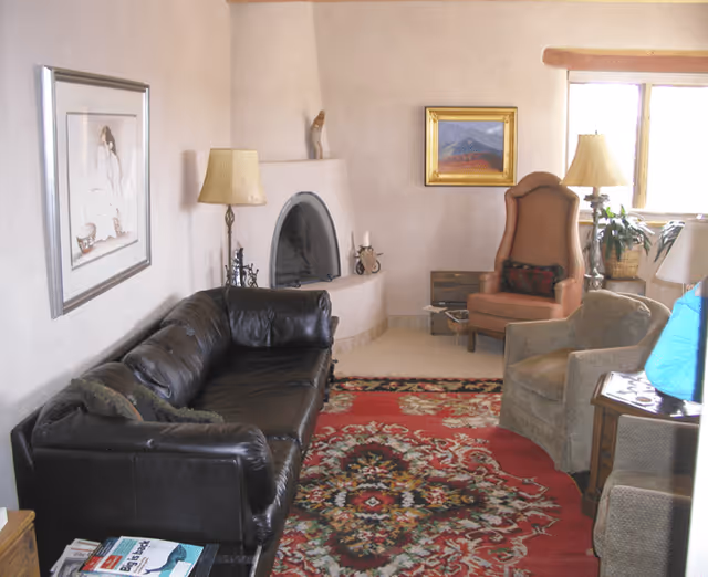 A cozy living room with a dark brown leather sofa, two armchairs, a colorful patterned rug, a corner fireplace, a floor lamp, framed artwork on the walls, and a window letting in natural light.