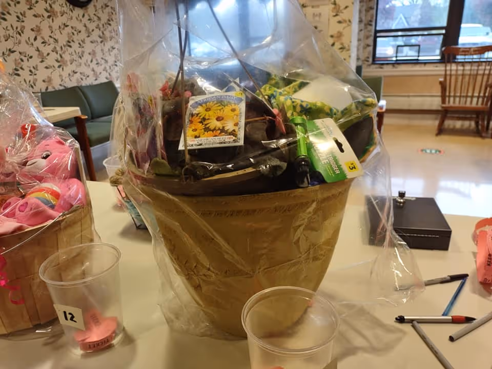 A large decorative pot wrapped in clear plastic containing various gardening items including a seed packet and a small green garden tool, placed on a table in a room with floral wallpaper, chairs, and windows in the background. Nearby on the table are two clear plastic cups with pink tickets inside and some pens.