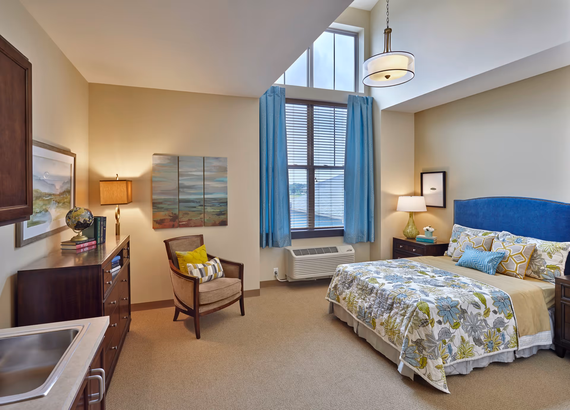 A cozy bedroom in a senior living facility with a bed featuring a blue upholstered headboard and floral bedding. The room has beige walls, a large window with blue curtains, a wooden dresser with a globe and books, a comfortable armchair with cushions, and a nightstand with a lamp and decor. There is also a small sink and countertop visible in the corner.