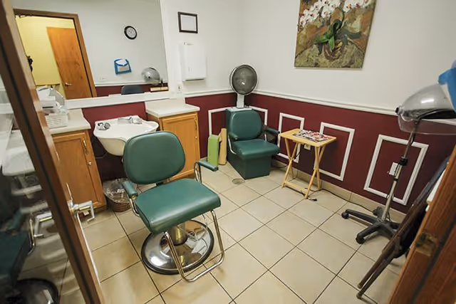 Interior of a small salon or hair care room with two green salon chairs, a white sink, a standing hair dryer, a small wooden table with magazines, and a painting on the wall. The room has tiled floors and white walls with a maroon wainscoting.