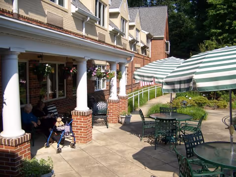 Outdoor patio with green metal tables and striped umbrellas beside a brick building with white columns, with two people seated and a walker nearby.