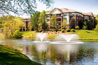 Brick multi-story senior living building beside a landscaped pond with two fountains and trees.