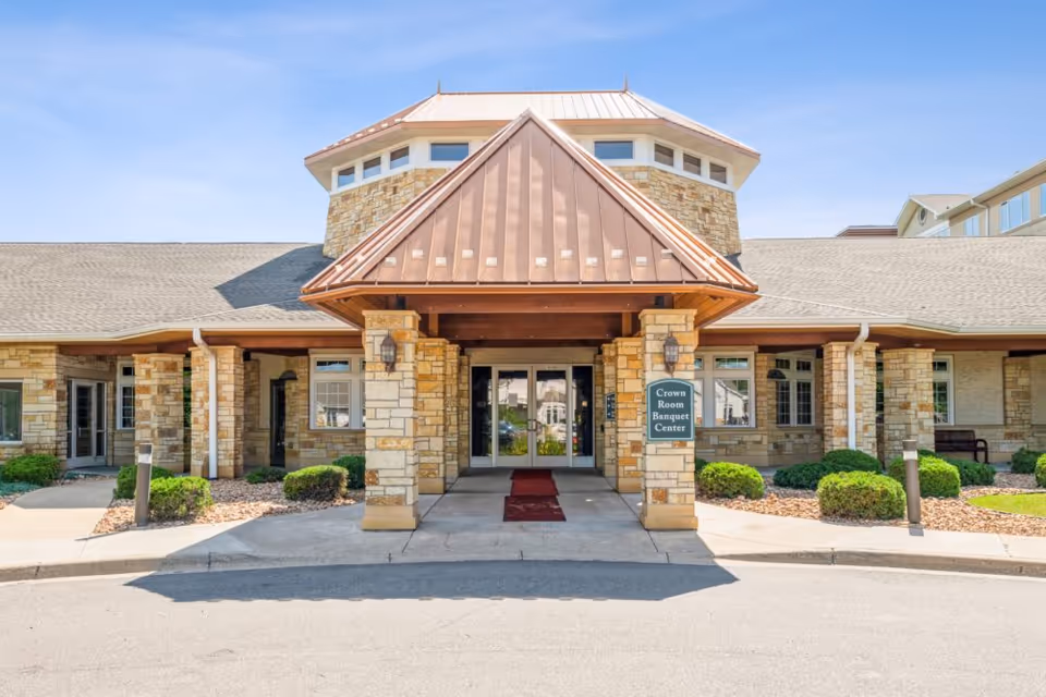Front exterior view of a senior living facility entrance with stone pillars and a copper-colored roof overhang. There is a sign that reads 'Crown Room Banquet Center' near the entrance, surrounded by neatly trimmed bushes and landscaping under a clear blue sky.