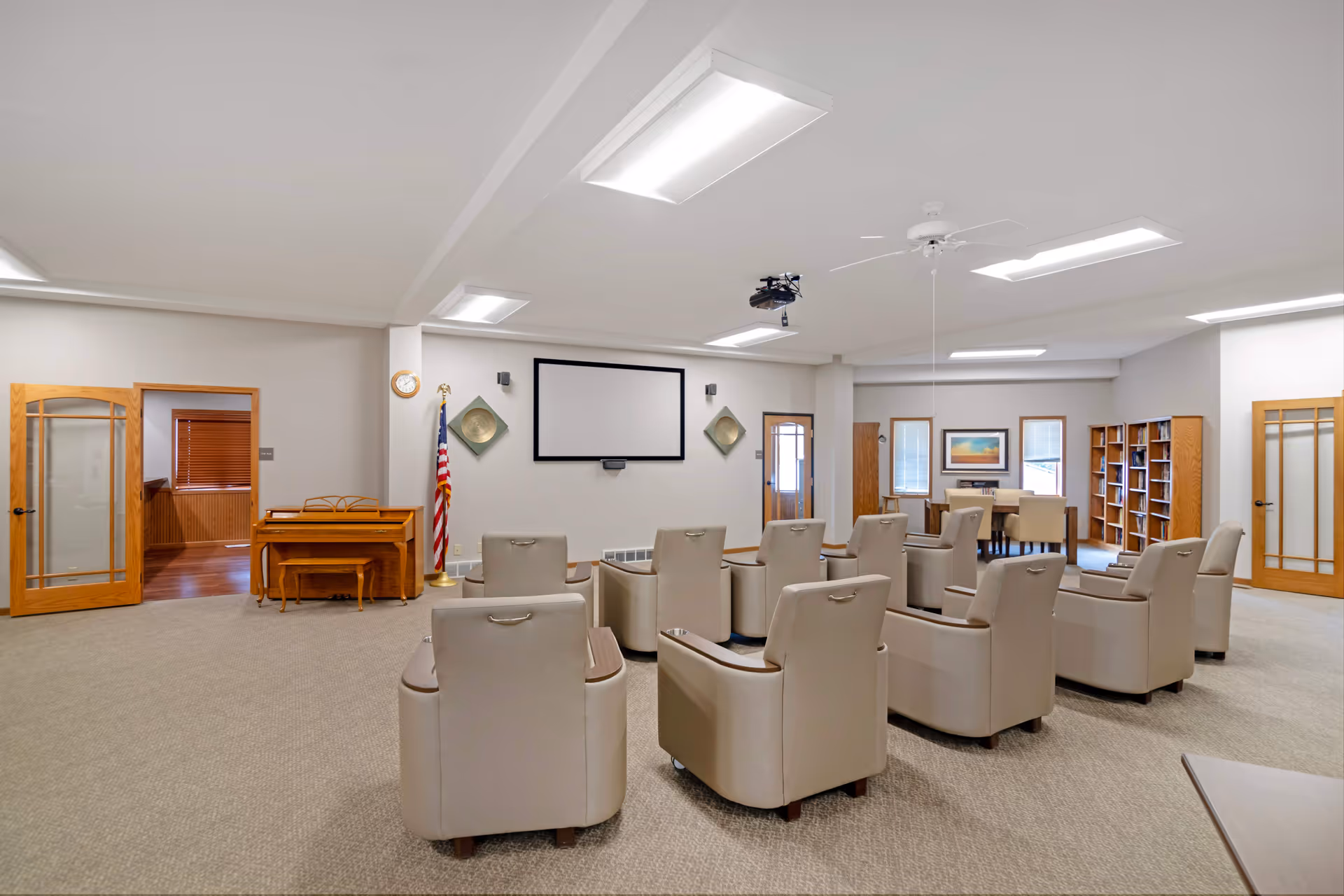 A spacious senior living common room with rows of beige armchairs facing a projector screen, a piano, and bookshelves.