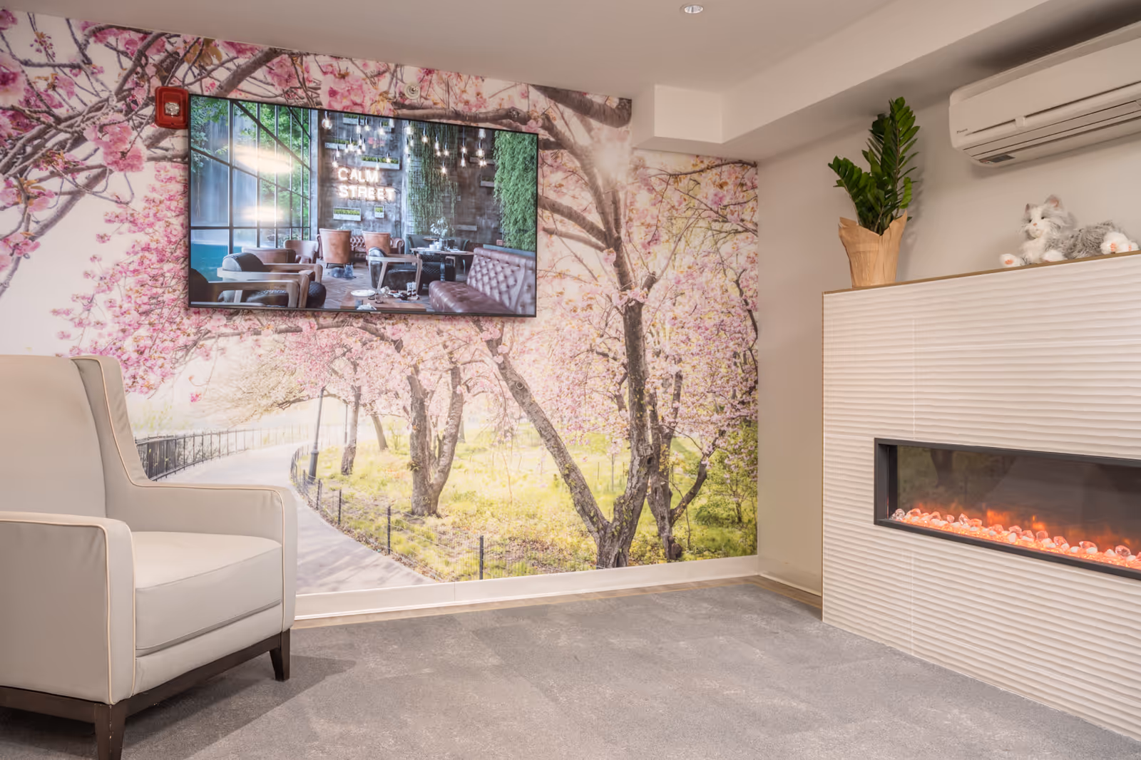 A cozy lounge area with a beige armchair on the left, a wall mural of a cherry blossom tree pathway, a flat-screen TV mounted on the wall displaying an image of a cafe with the text 'CALM STREET', and a modern electric fireplace on the right with a potted plant and a plush cat toy on top.
