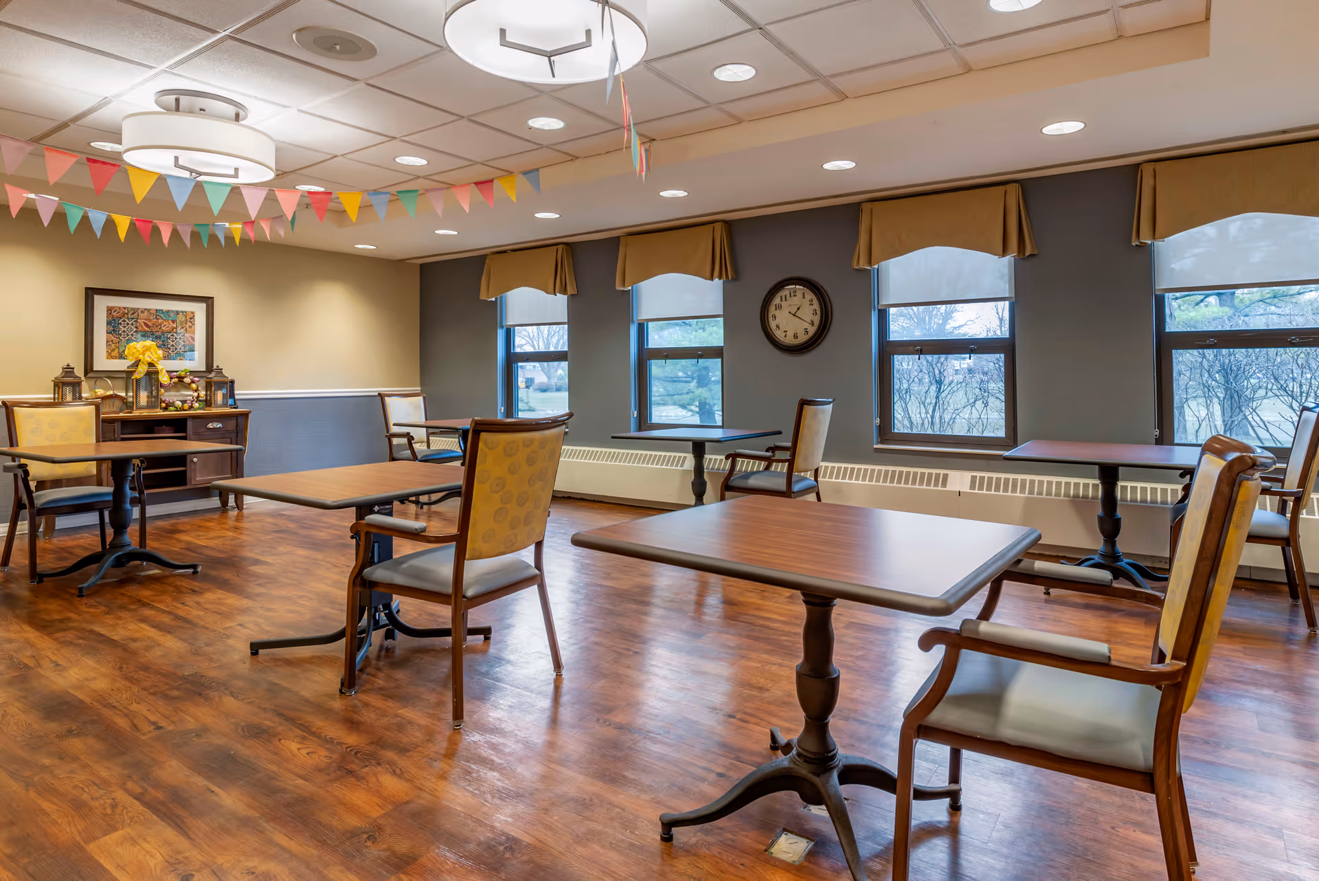 A dining room with wooden floors, several tables and chairs spaced apart, large windows with beige valances, a wall clock, and colorful triangular pennant banners hanging from the ceiling. A sideboard with decorative items is against one wall.