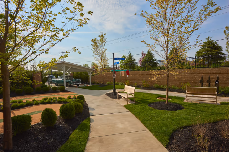 A landscaped outdoor area with a concrete walkway, green grass, small trees, and bushes. There are two wooden benches along the path and a covered parking area with a vintage green car parked underneath. A street sign is visible near the benches, and a retaining wall with fencing is in the background under a partly cloudy sky.