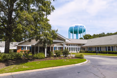 Exterior view of Churchland Nursing Home building with a curved driveway, green lawn, bushes, and a large tree. A blue water tower with the word 'PORTSMOUTH' is visible in the background under a partly cloudy sky.