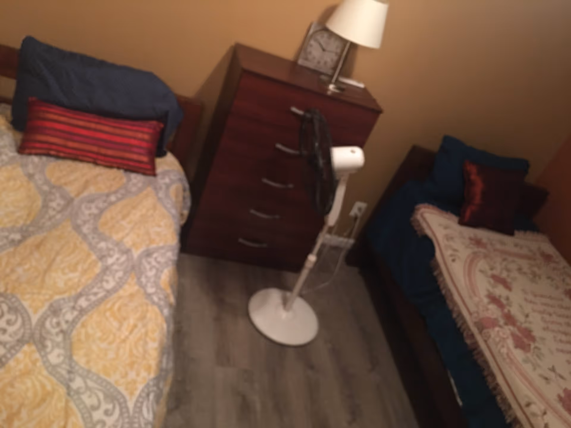 A small bedroom with two single beds separated by a wooden dresser. Each bed has pillows and patterned blankets. A white standing fan is placed in front of the dresser. A table lamp and a clock are on top of the dresser. The room has wooden flooring and beige walls.