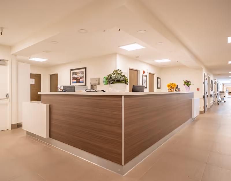 A modern reception desk in a well-lit hallway of a senior living facility. The desk has a wood finish with a white countertop and is decorated with a potted plant and flowers. The hallway features beige tiled floors, white walls, several closed wooden doors, framed artwork, and ceiling lights.