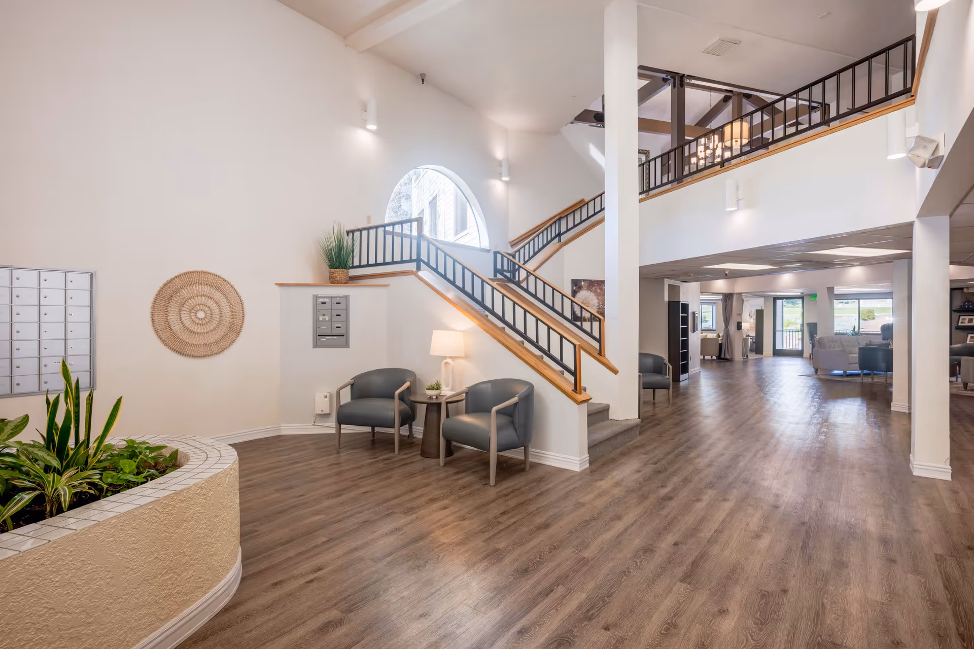 Spacious and well-lit interior common area of a senior living facility featuring a staircase with black railings and wooden handrails, two gray chairs with a small round table and lamp between them, a large circular window, indoor plants in a tiled planter, and wood-style flooring extending throughout the space.