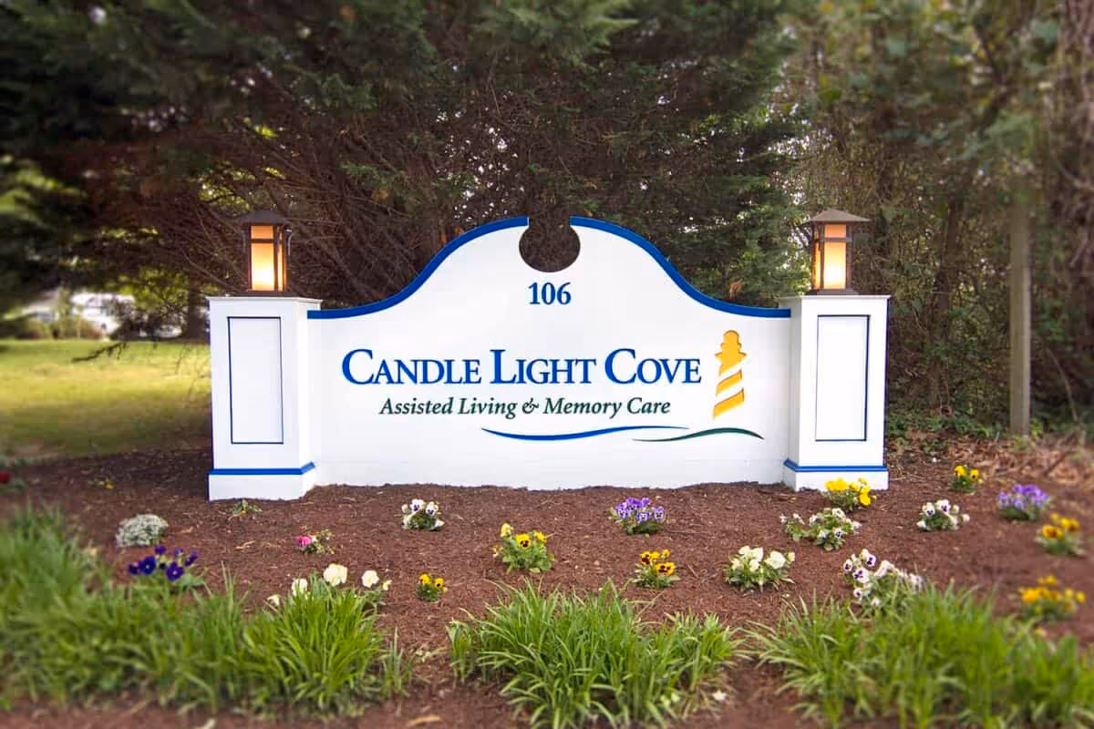 A white sign with blue trim and two lanterns on either side, displaying the text '106 Candle Light Cove Assisted Living & Memory Care' with a yellow lighthouse graphic. The sign is surrounded by mulch, small flowering plants, and greenery, with trees in the background.