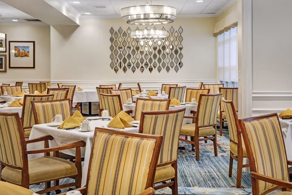 A dining room with multiple tables covered in white tablecloths, each set with yellow folded napkins, white cups, and silverware. The chairs have wooden frames with striped yellow and beige upholstery. The room features a patterned carpet, two modern circular chandeliers, a decorative wall mirror with a wavy design, and framed artwork on the wall.