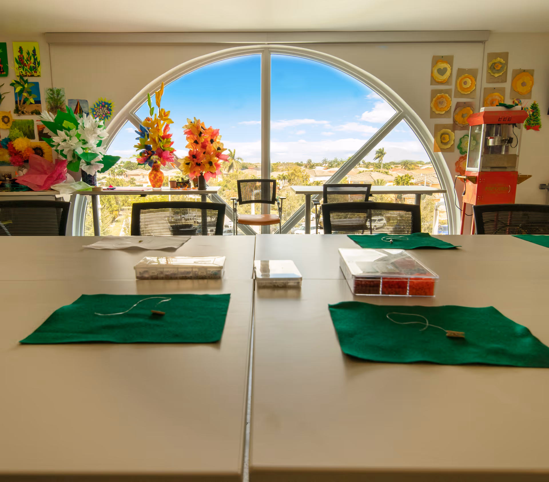 A bright activity room with a large arched window showing a sunny outdoor view. The room has tables with green mats and craft supplies, chairs, colorful paper flower decorations, and a popcorn machine on the right side.