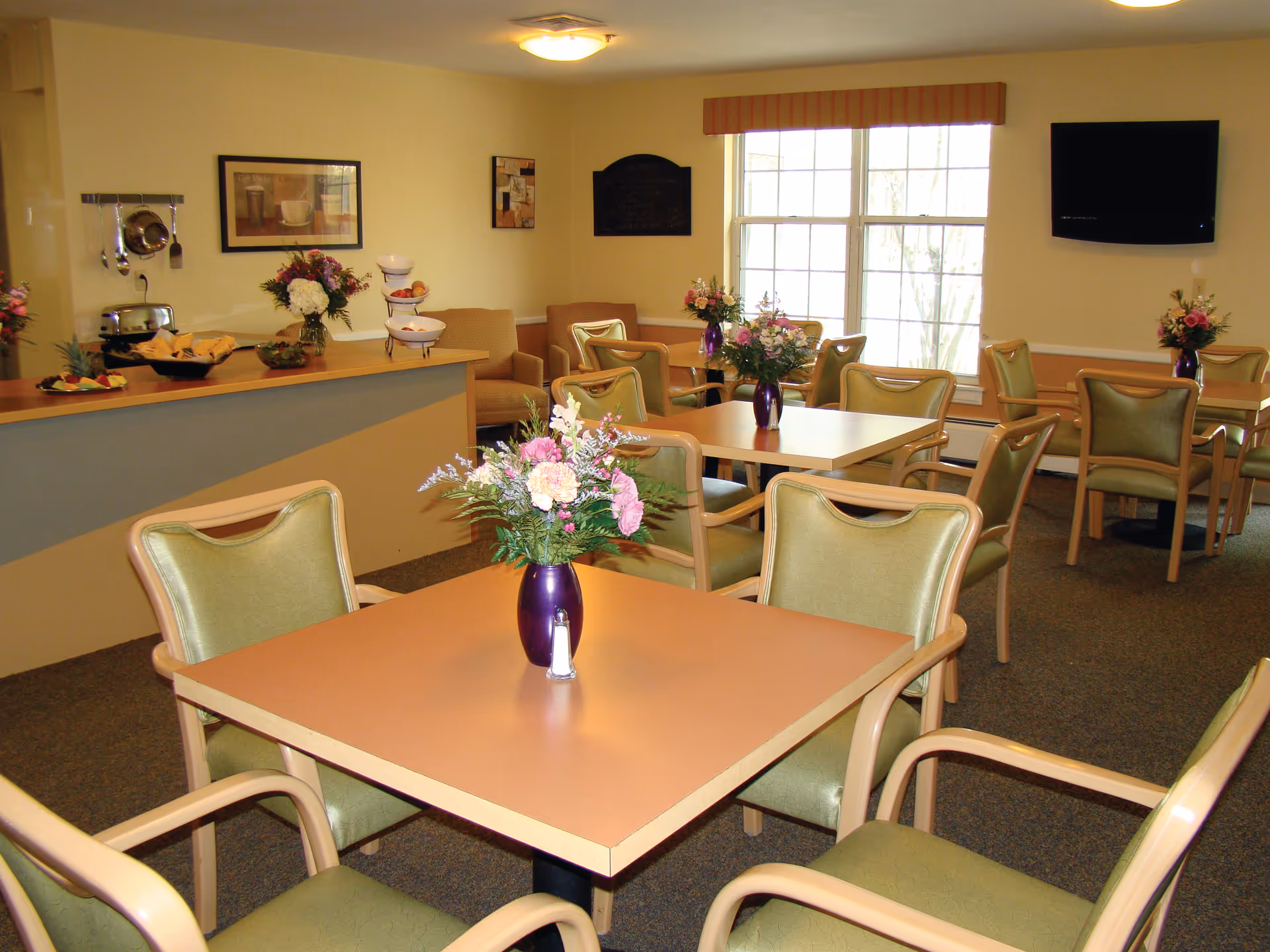A dining room with several square tables, each surrounded by four green cushioned chairs. Each table has a purple vase with a bouquet of flowers. There is a counter with fruit and a toaster on it, and a large window with a striped valance letting in natural light. A flat-screen TV is mounted on the wall.