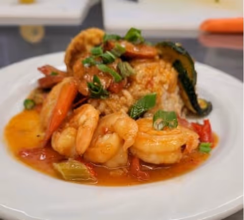 A close-up of a plate of shrimp cooked in a reddish sauce with chopped green onions on top, served with rice and vegetables including zucchini and red bell peppers.